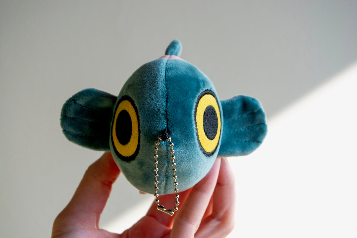 Hand holding a small blue plush fish keychain with large yellow eyes and soft mochi fabric texture