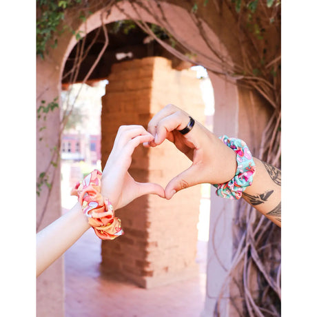 Two hands wearing colorful Capybara Hair Scrunchie forming a heart shape outdoors near archway.