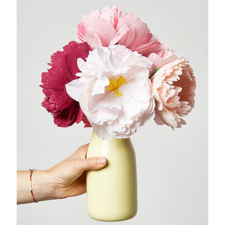 Hand holding a yellow vase filled with handmade paper flowers in soft pink, white, and deep red shades against a white background