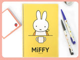Yellow spiral notebook with a simple white bunny illustration holding a cup and two decorated pens on the side