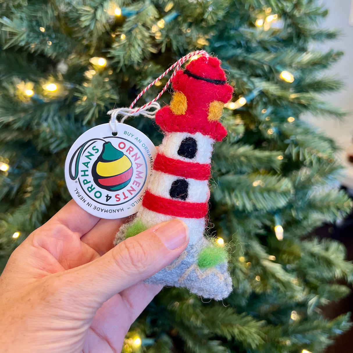 Handmade red and white felt lighthouse ornament held in hand with Christmas tree lights in background