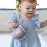 Toddler girl in light blue dress holding a pink sippy cup with gray handles and "Spill The Tea" text