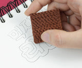 Hand holding a brown eraser shaped like a block of ramen noodles erasing pencil drawing on a spiral notebook page