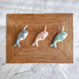 Three hand-felted narwhal ornaments in blue, pink, and teal on a woven placemat background