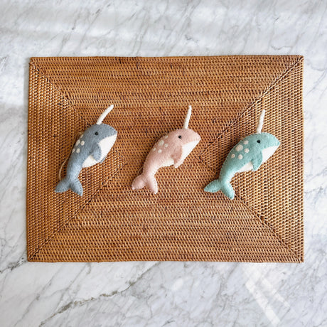 Three hand-felted narwhal ornaments in blue, pink, and teal on a woven placemat background