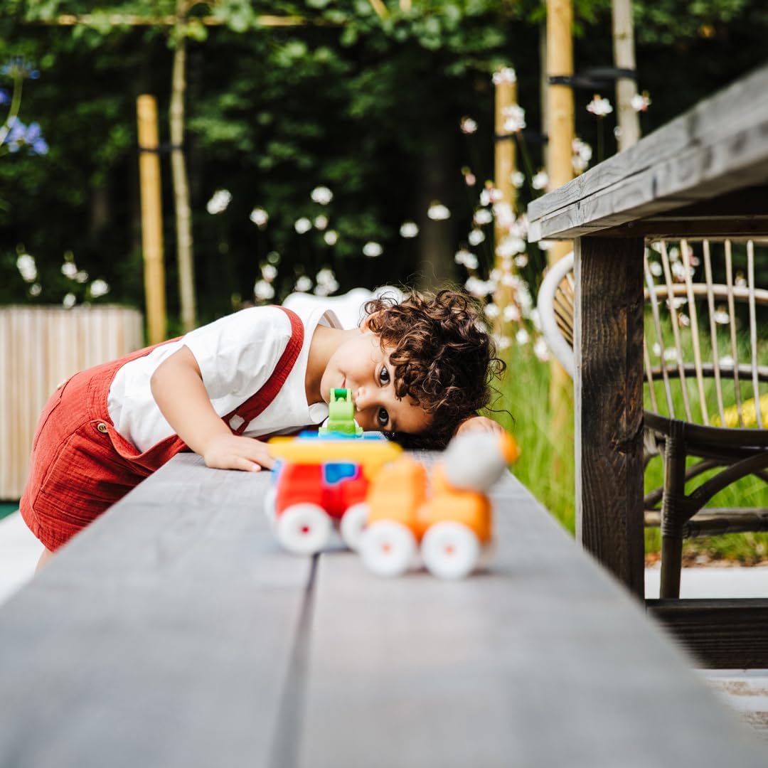 Young child with curly hair leaning on wooden table playing with colorful toy train outdoors in garden setting