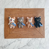 Four handmade felt cat ornaments in different colors each holding a small blue fish on a woven mat