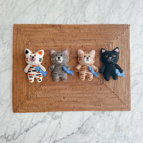 Four handmade felt cat ornaments in different colors each holding a small blue fish on a woven mat