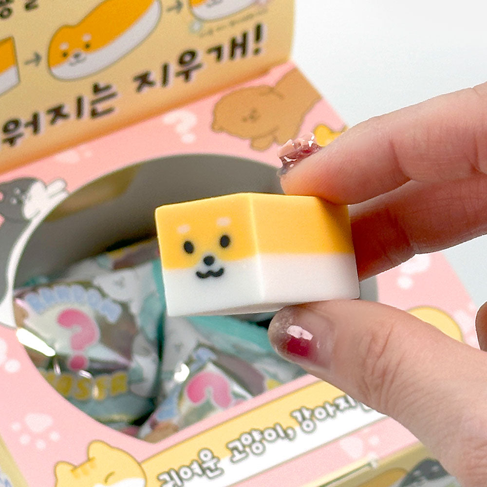 Hand holding a small yellow and white dog-shaped eraser with a smiling face above a box of pet eraser blind bags