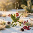 Green felt cricket ornament wearing a red Santa hat hanging on snow with pine cones and berries surrounding it