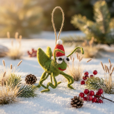 Green felt cricket ornament wearing a red Santa hat hanging on snow with pine cones and berries surrounding it