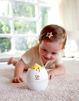 Baby lying on carpet smiling at a yellow chick popping out of a white egg toy on the floor