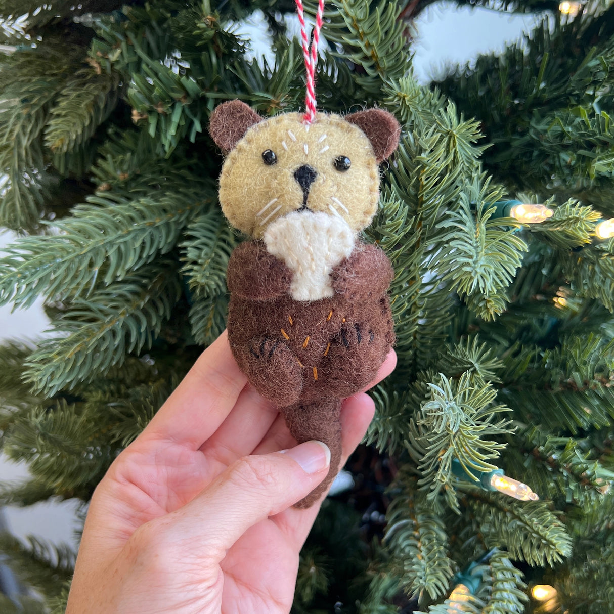 Handmade felt otter ornament in brown and tan wool hanging on Christmas tree branch outdoors