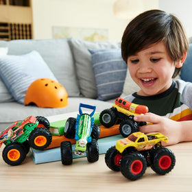 Child happily playing with Hot Wheels Monster Truck 4pk featuring colorful 1:64 scale trucks with giant wheels on a wooden table.