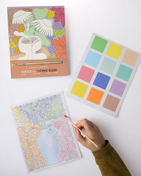 Person painting floral line art on canvas sheet from Water Painting Book set with color palette guide nearby on white surface