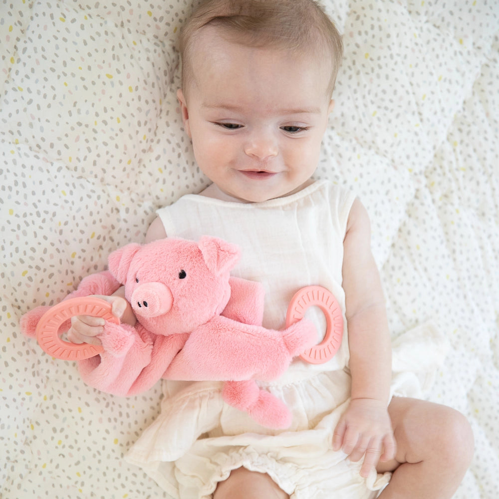 Infant holding a soft pink pig plush with silicone teething rings while lying on a patterned blanket