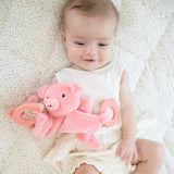 Infant holding a soft pink pig plush with silicone teething rings while lying on a patterned blanket