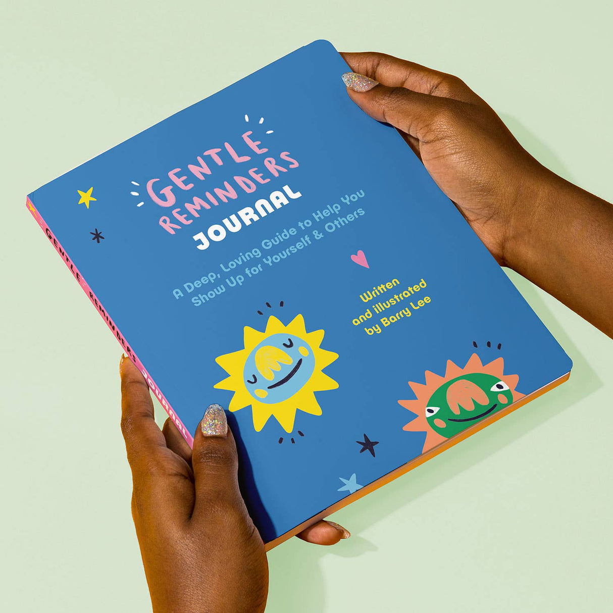 Hands holding a colorful Gentle Reminders Guided Journal with playful sun illustrations on the cover against a pale background