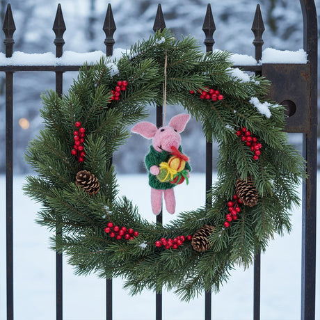 Handfelted pig ornament wearing a green cardigan holding gifts, hanging in a festive pine and red berry wreath outdoors in snow.