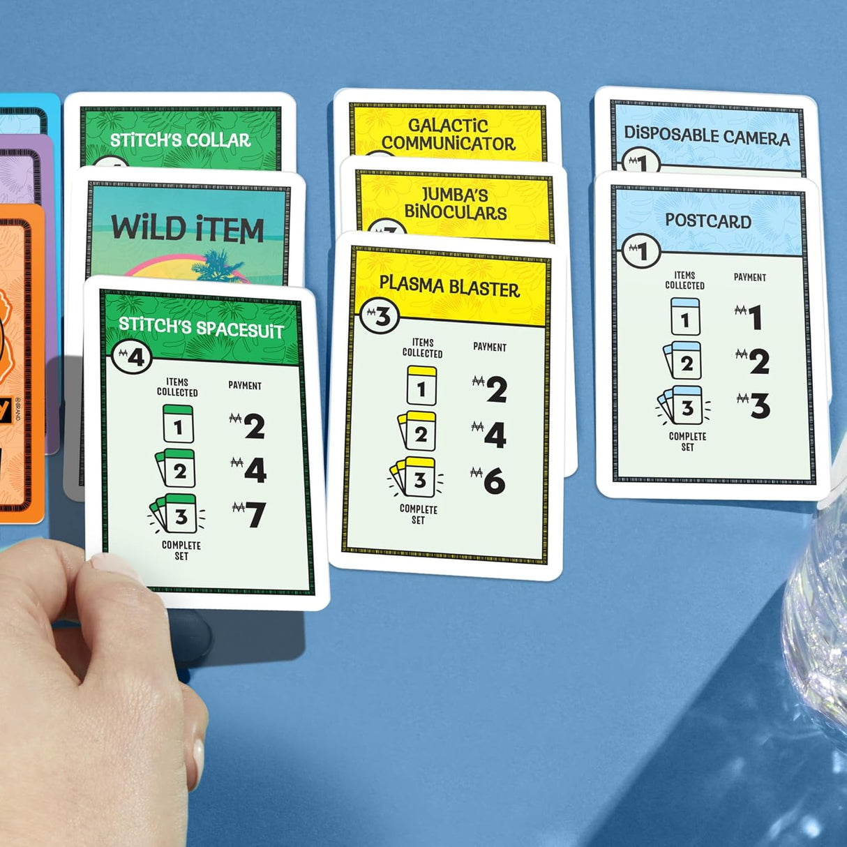 Hand holding cards from the Monopoly Deal Disney Stitch edition showing items like Stitch's spacesuit and plasma blaster on a blue background