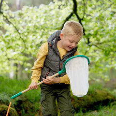 Young boy exploring outdoors with a green and white bug-catching net in a leafy forest