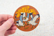 Hand holding a round sticker with three seagulls and the word gather in bright yellow script on an orange background