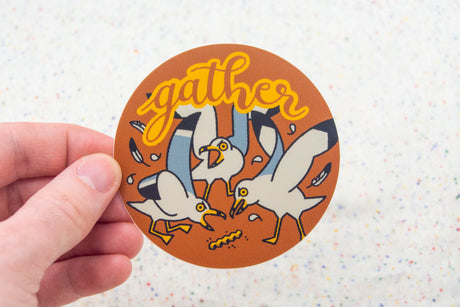 Hand holding a round sticker with three seagulls and the word gather in bright yellow script on an orange background