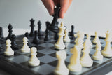 Close-up of a hand moving a black chess piece on a magnetic travel chessboard with white pieces in focus