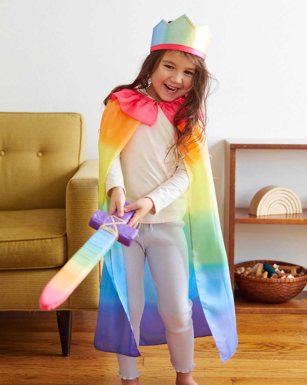 Smiling child wearing a rainbow cape and matching crown holding a soft fabric sword indoors