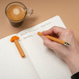 Erasable Gel Pen | Croissant orange pen with croissant topper shown writing in a lined notebook beside coffee