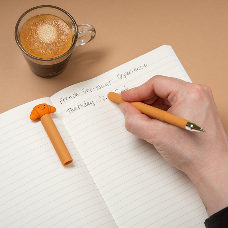 Erasable Gel Pen | Croissant orange pen with croissant topper shown writing in a lined notebook beside coffee
