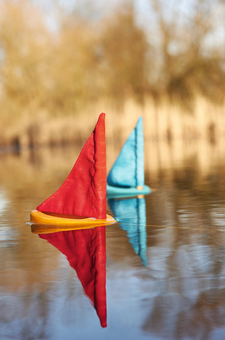 Two handcrafted wooden sailboats with red and turquoise fabric sails floating on calm water outdoors