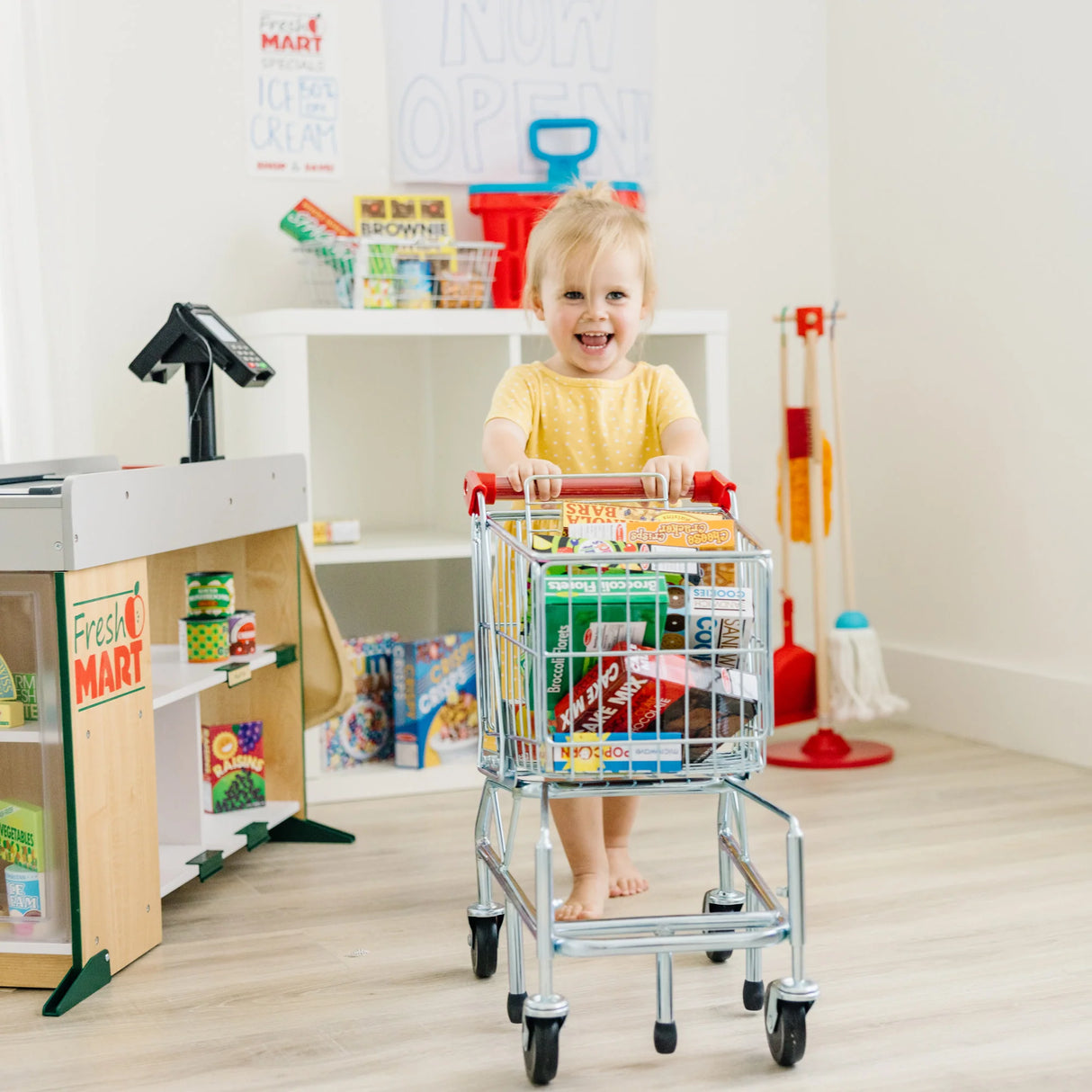 Toddler in yellow outfit pushing a toy shopping cart filled with play food boxes in a bright playroom