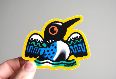 Hand holding a colorful 3-inch weatherproof vinyl sticker of a stylized loon bird with bold patterns and a yellow border