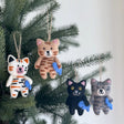 Handmade felt cat Christmas ornaments in various colors each holding a small blue fish hanging on a pine tree branch
