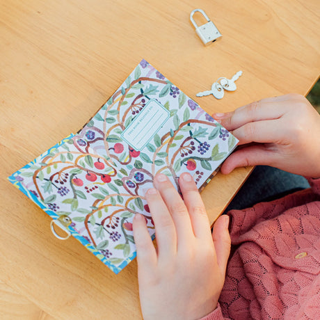 Very Small Journal | Gemma open showing floral illustration pages with hands, metal lock and keys on wooden table.