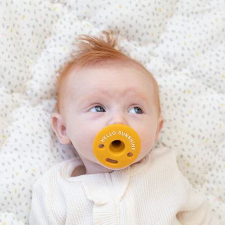 Baby lying down using a bright yellow pacifier with "Hello Sunshine" text, Pacifier | Hello Sunshine product.