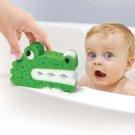 Hand holding Bath Biters Croc sponge with soap bubbles near surprised baby in bathtub.