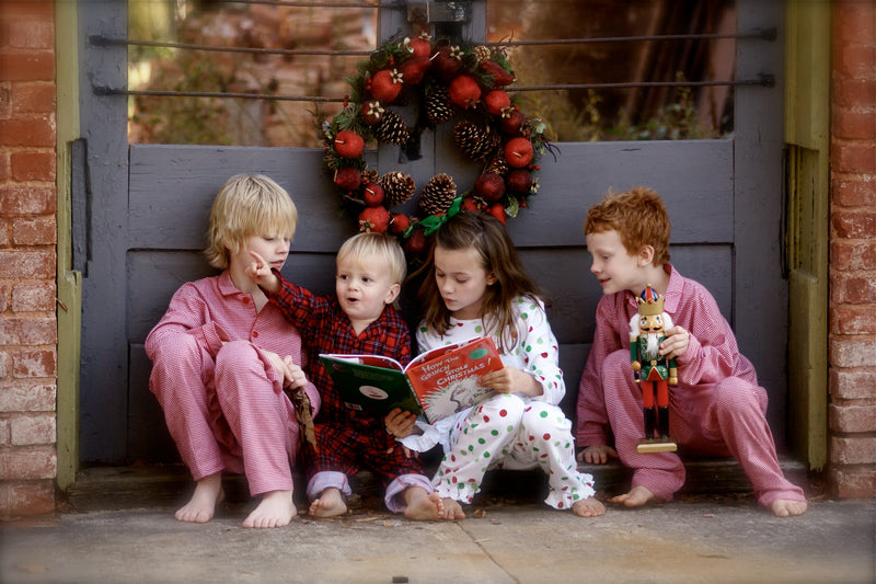 Four children in holiday pajamas sitting by a door with a festive wreath reading a Christmas book together