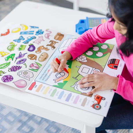 Child coloring a farm scene with crayons in the Colors & Shapes Activity Pad on a white table.