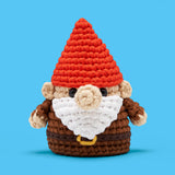 Crocheted gnome with red hat, white beard, and round nose standing against a blue background