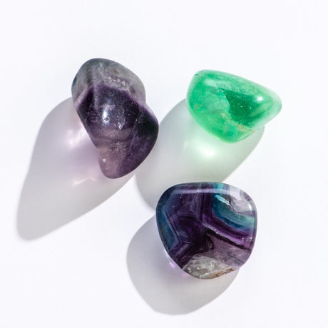 Three polished tumbled rainbow fluorite stones in green, purple, and blue shades on a white background.