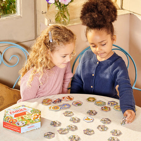 Two children playing Cottage Fairies Memory & Matching Game with illustrated hexagonal tiles on a table.