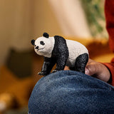 Textured figurine of a standing giant panda held above a person's denim-clad knee in a warm indoor setting