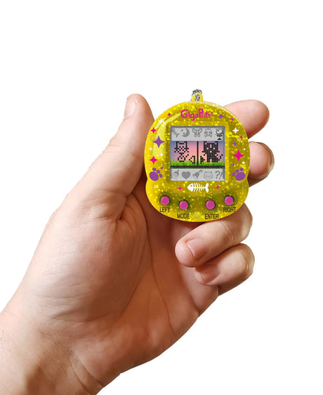 Hand holding a yellow GigaPets Starcat Compukitty virtual pet with pixelated cat display and control buttons.