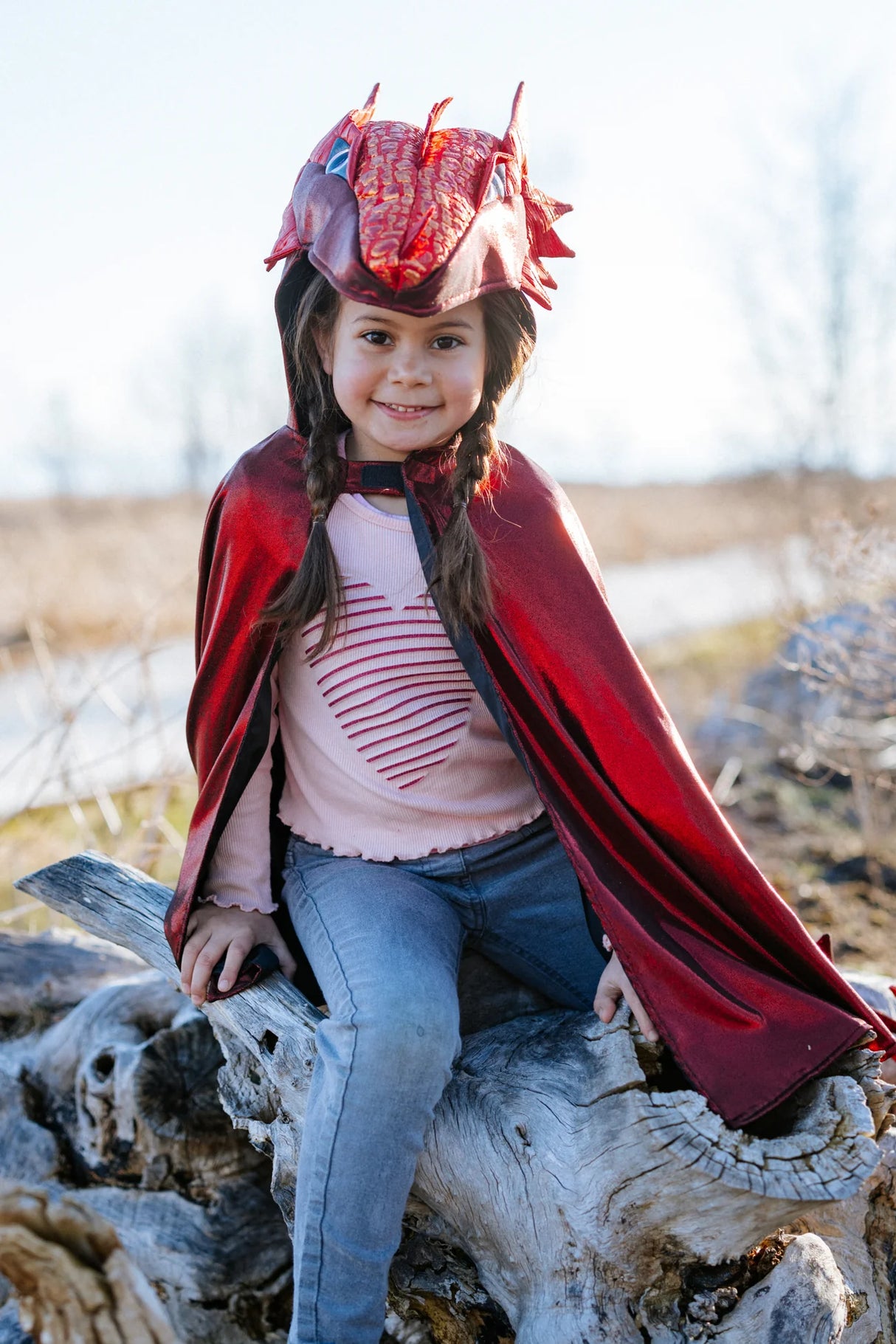 Child wearing Ruby the Metallic Dragon Cape Size 5-6 with shiny red dragon hood sitting on a log outdoors
