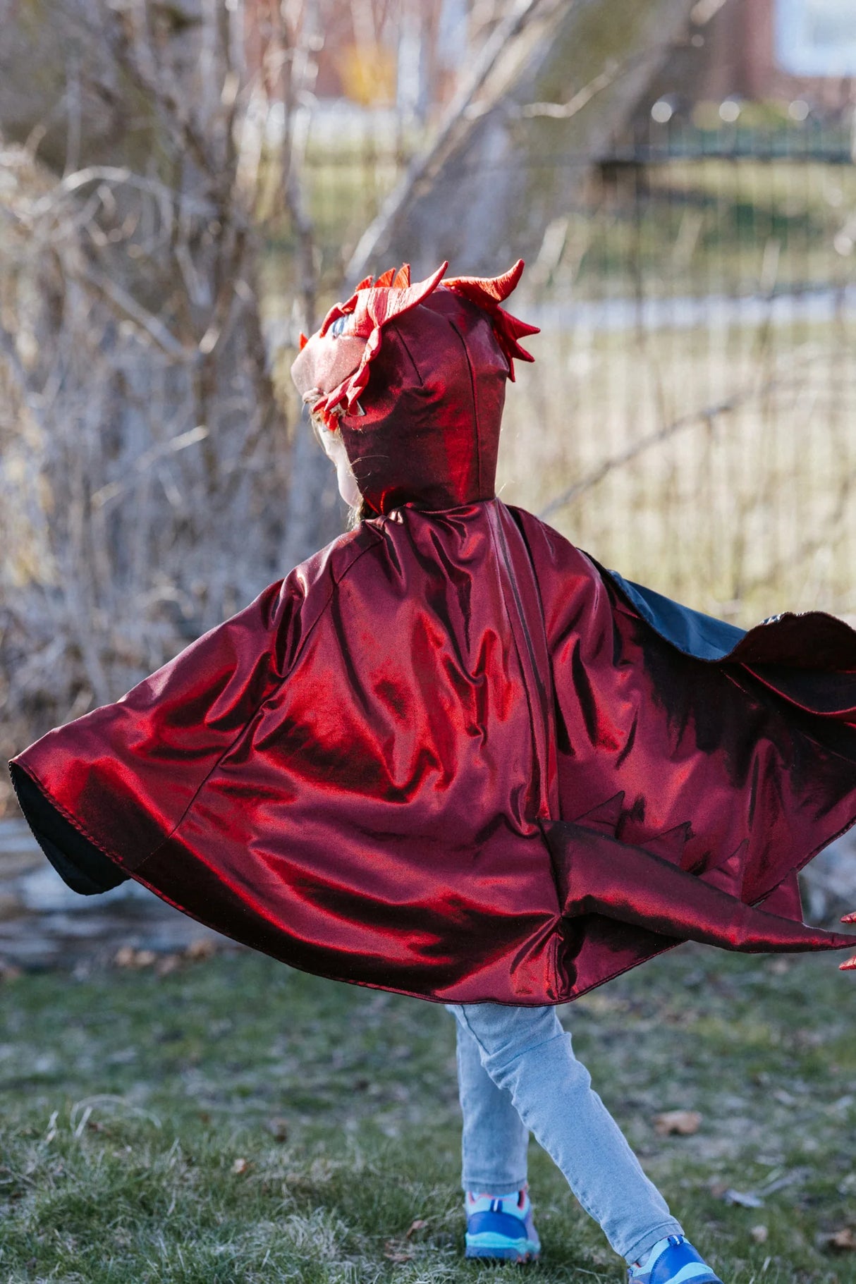 Ruby the Metallic Dragon Cape Size 5-6 shown worn outdoors, shimmering red with dragon head and scale details.