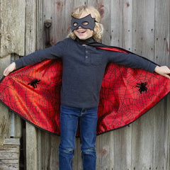 Child wearing a black eye mask and red spiderweb cape with arms outstretched against a wooden fence