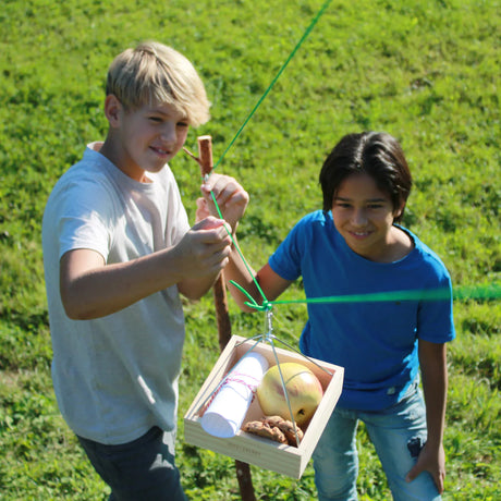 Two boys using the Huckleberry Cable Transport to send a wooden tray with items across an outdoor rope line.