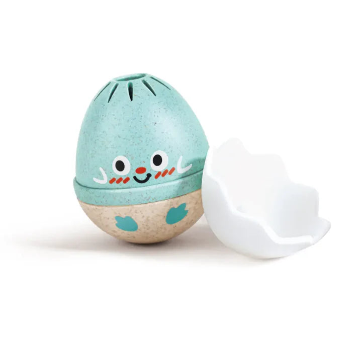 Blue and beige egg-shaped baby rattle with a smiling face and a white cracked eggshell beside it on a white background